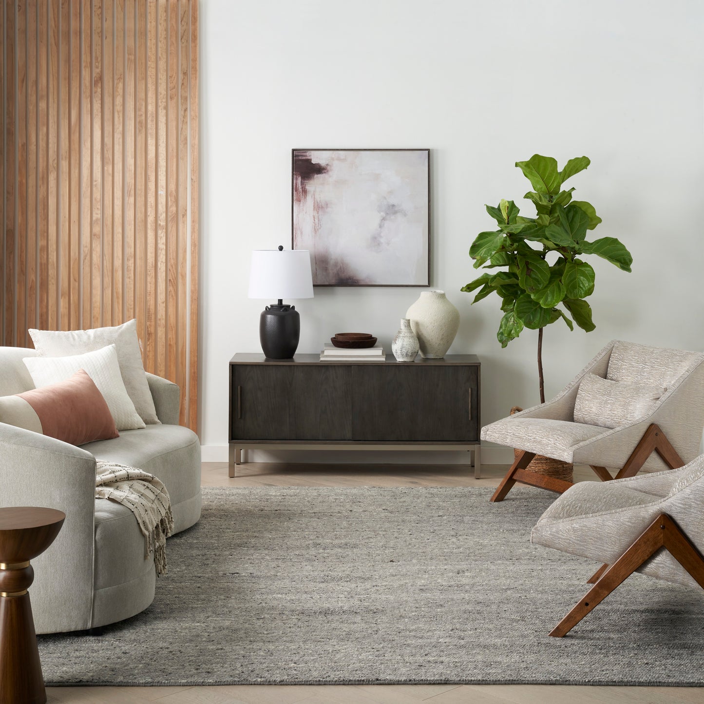 Modern living room with dark wood sideboard, abstract art, vases, large green plant, beige lounge chairs & gray rug.
