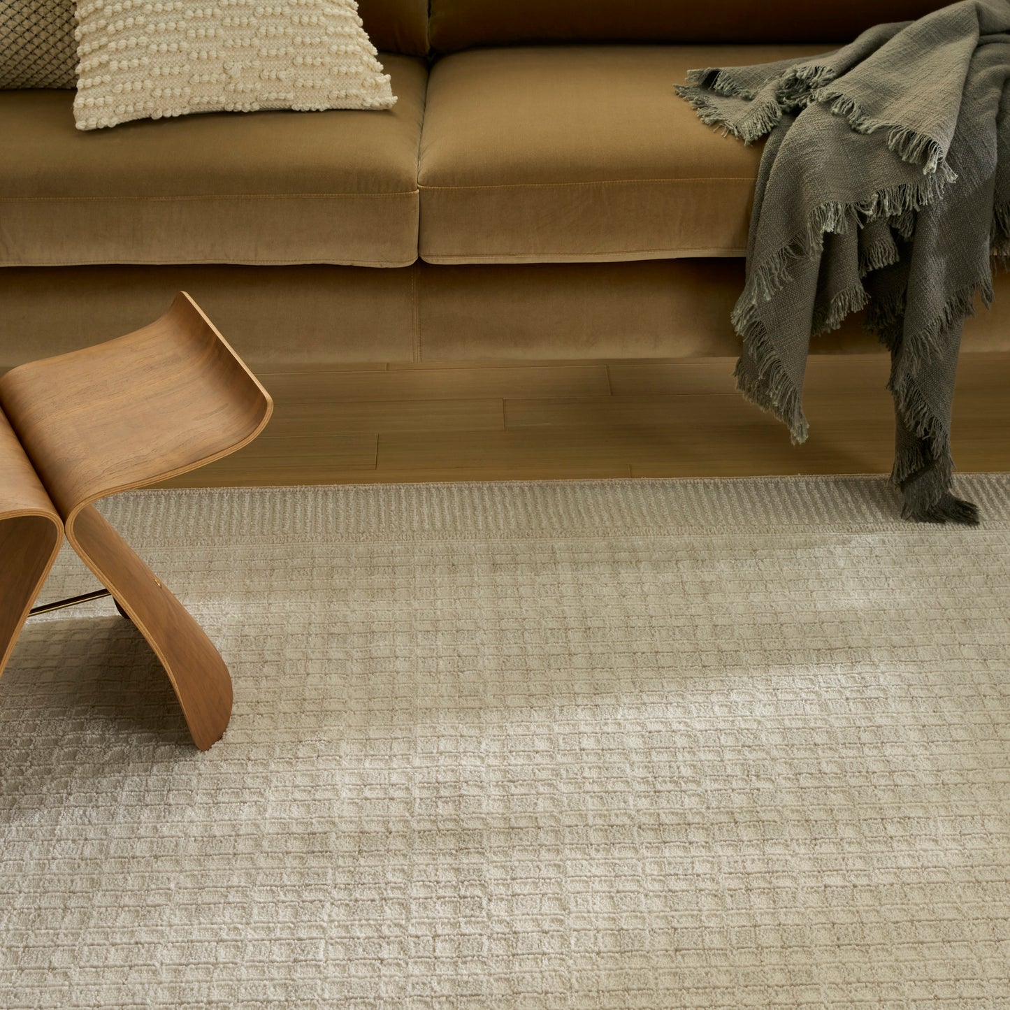 Beige mid-century sofa with textured cream pillow, gray fringe throw, curved wooden chair, and light grid rug on wood floor