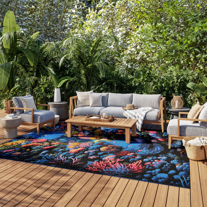 Outdoor lounge with light gray cushioned wooden sofa and chairs, vibrant coral-pattern rug, coffee table, and lush tropical greenery.