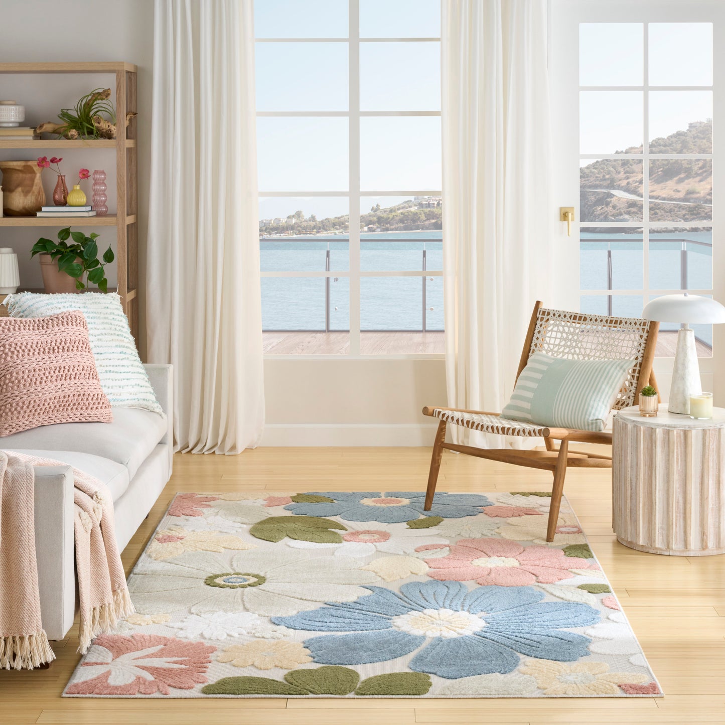 Pastel floral area rug in a coastal living room with light wood floor, white sofa, wicker chair, and sea-view windows.