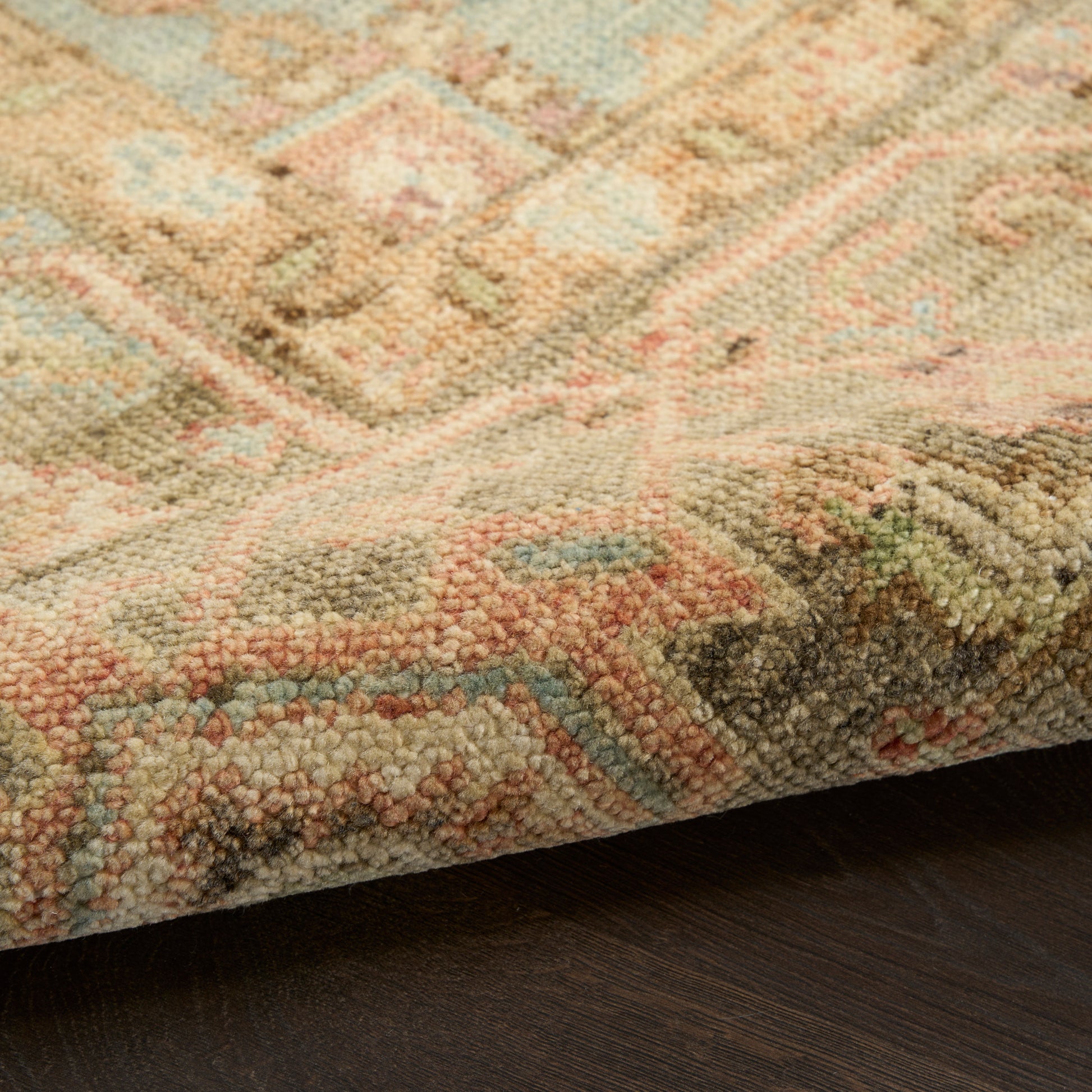 Pastel oriental rug with pink, blue, beige and green geometric motifs; thick plush pile, edge folded over dark wood floor.