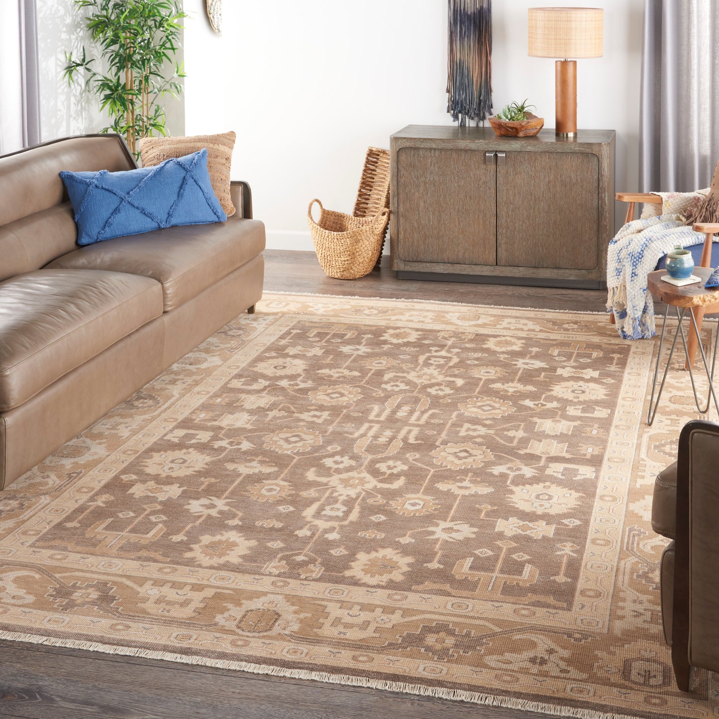 Brown leather sofa, blue cushions, beige geometric area rug, wooden cabinet, plant, lamp and woven basket in a living room.