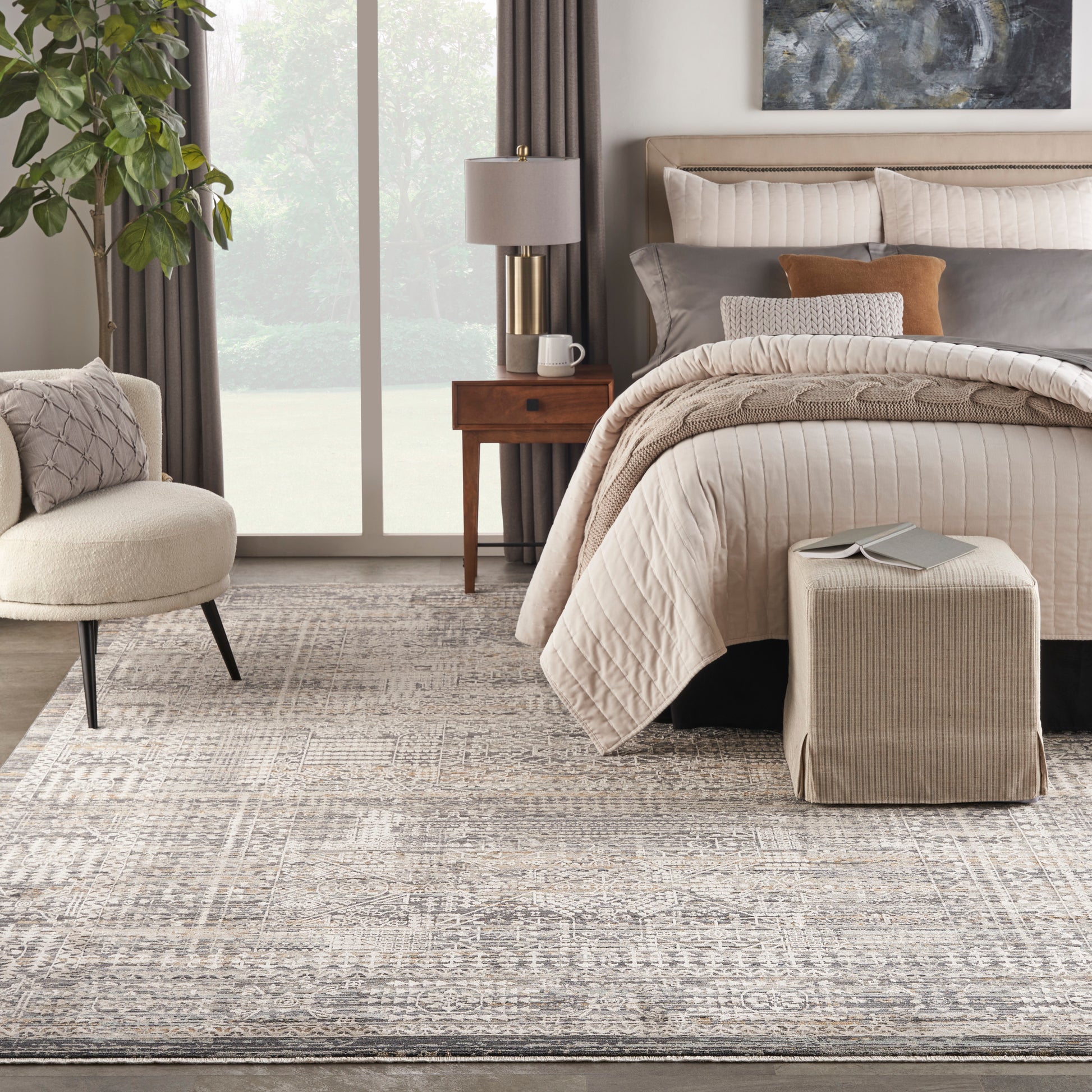 Neutral-toned bedroom with quilted beige bedding, fabric bench ottoman, textured rug, wooden nightstand, and greenery.