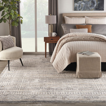 Neutral-toned bedroom with quilted beige bedding, fabric bench ottoman, textured rug, wooden nightstand, and greenery.