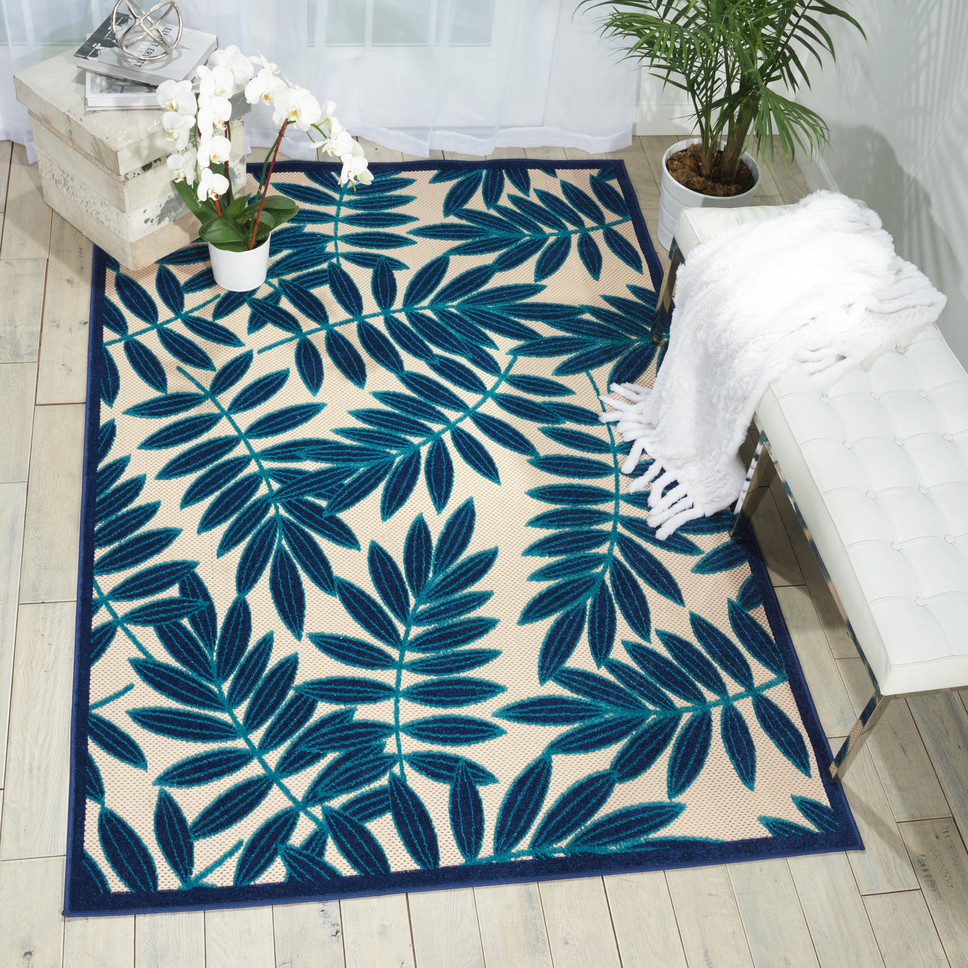 Teal & navy palm-leaf patterned area rug on light wood floor with a white chair, throw, and potted orchid.