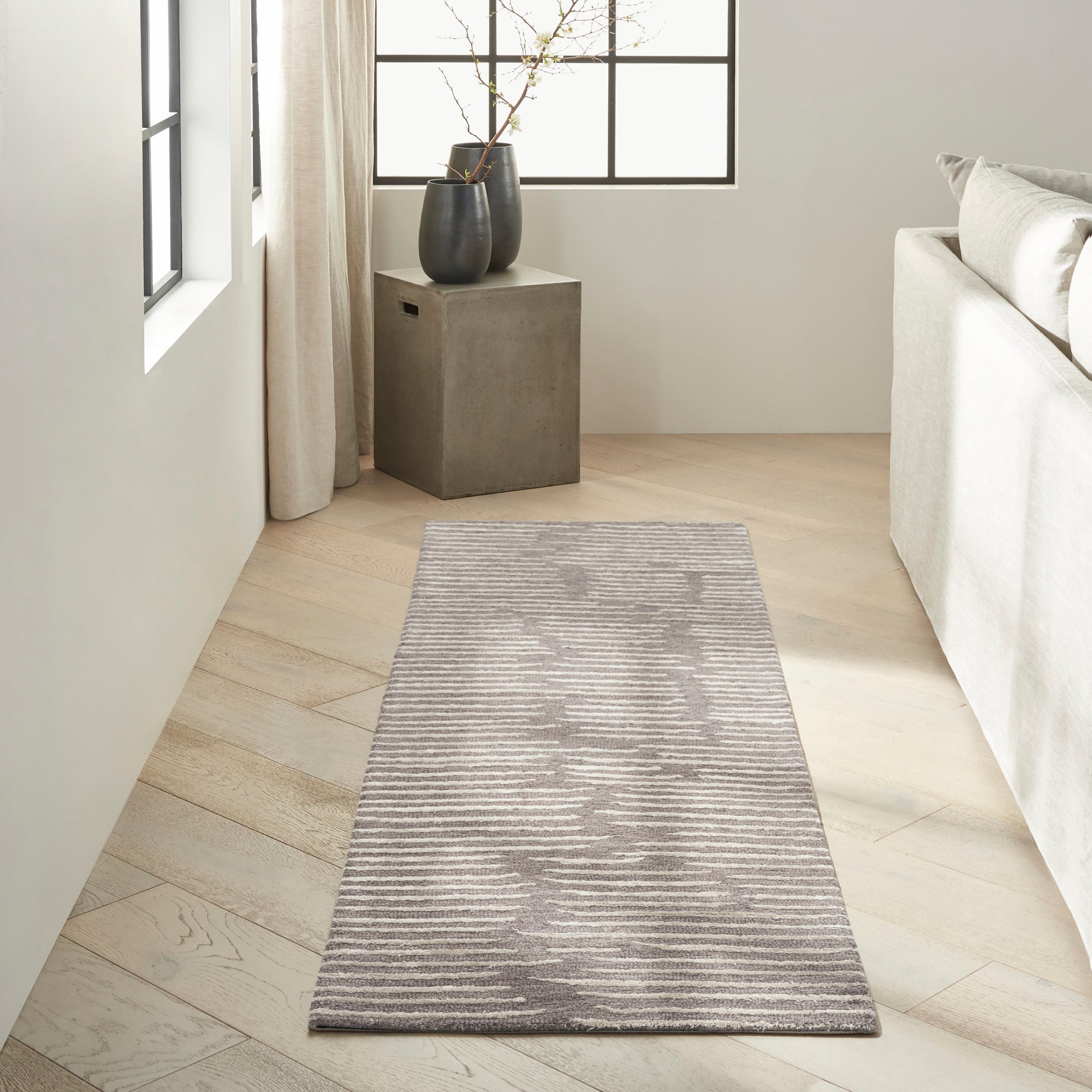 Gray striped runner rug on light oak floor in a bright living room, beige sofa nearby, concrete side table with black vases.