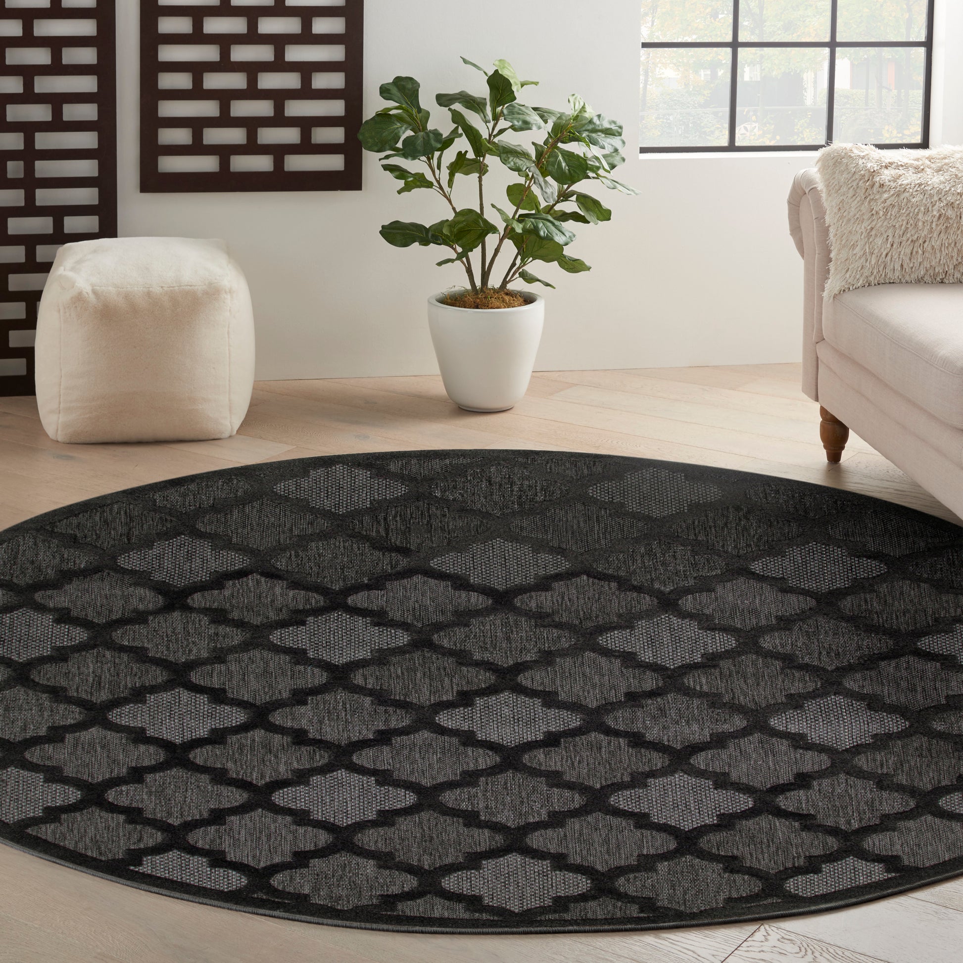 Round black rug with gray trellis lattice pattern in a bright living room with a beige sofa, cream pouf, and potted plant.