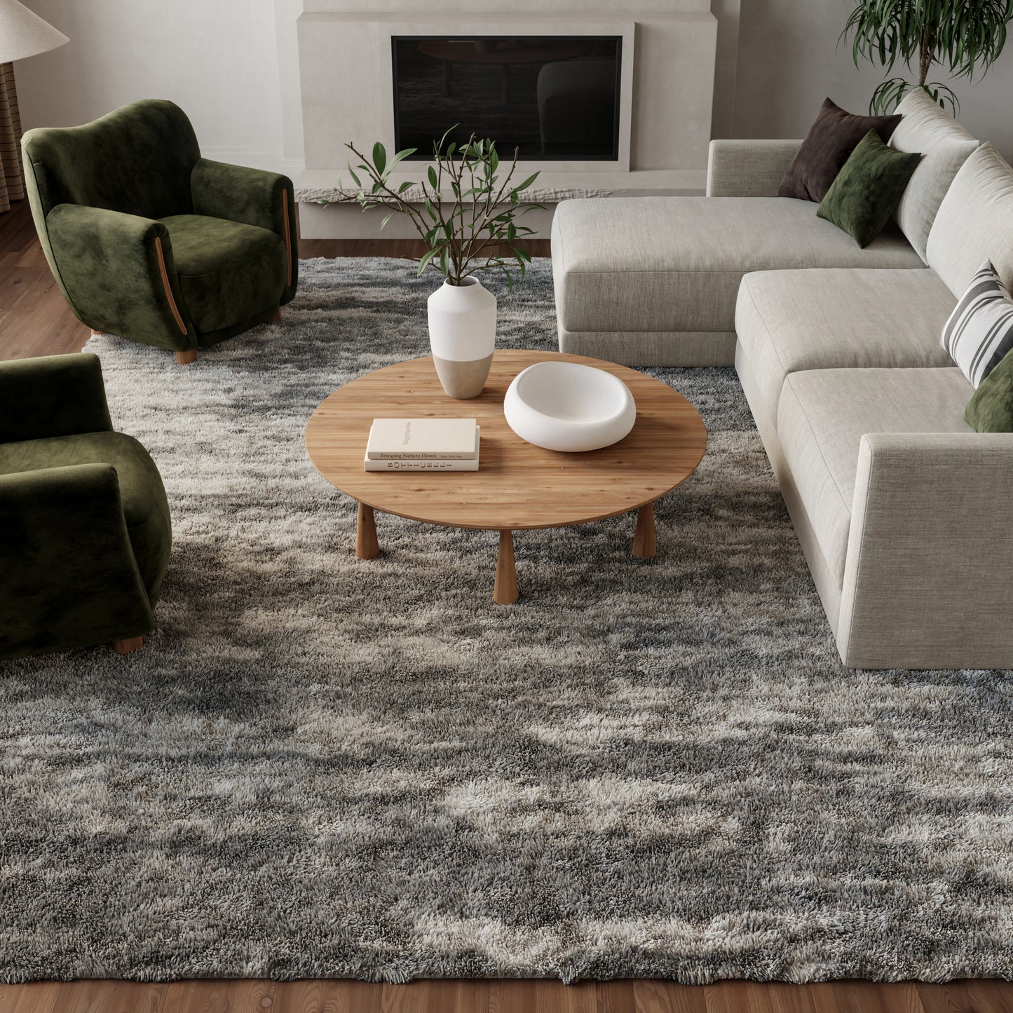 Round wooden coffee table with vase and bowl in a beige sectional living room with green velvet armchairs and a gray rug.