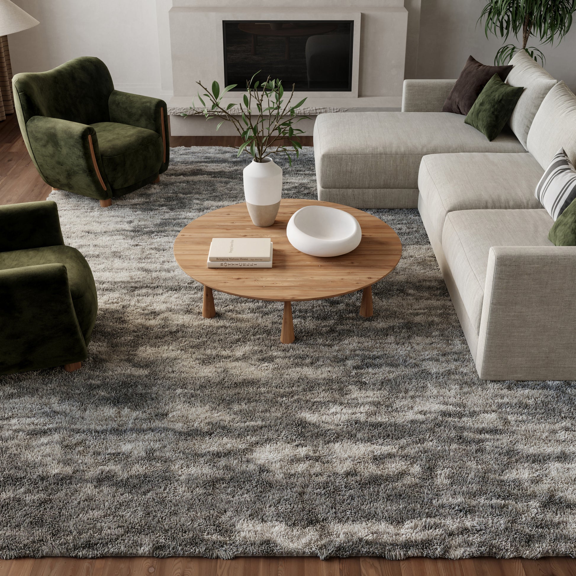 Round wooden coffee table with vase and bowl in a beige sectional living room with green velvet armchairs and a gray rug.