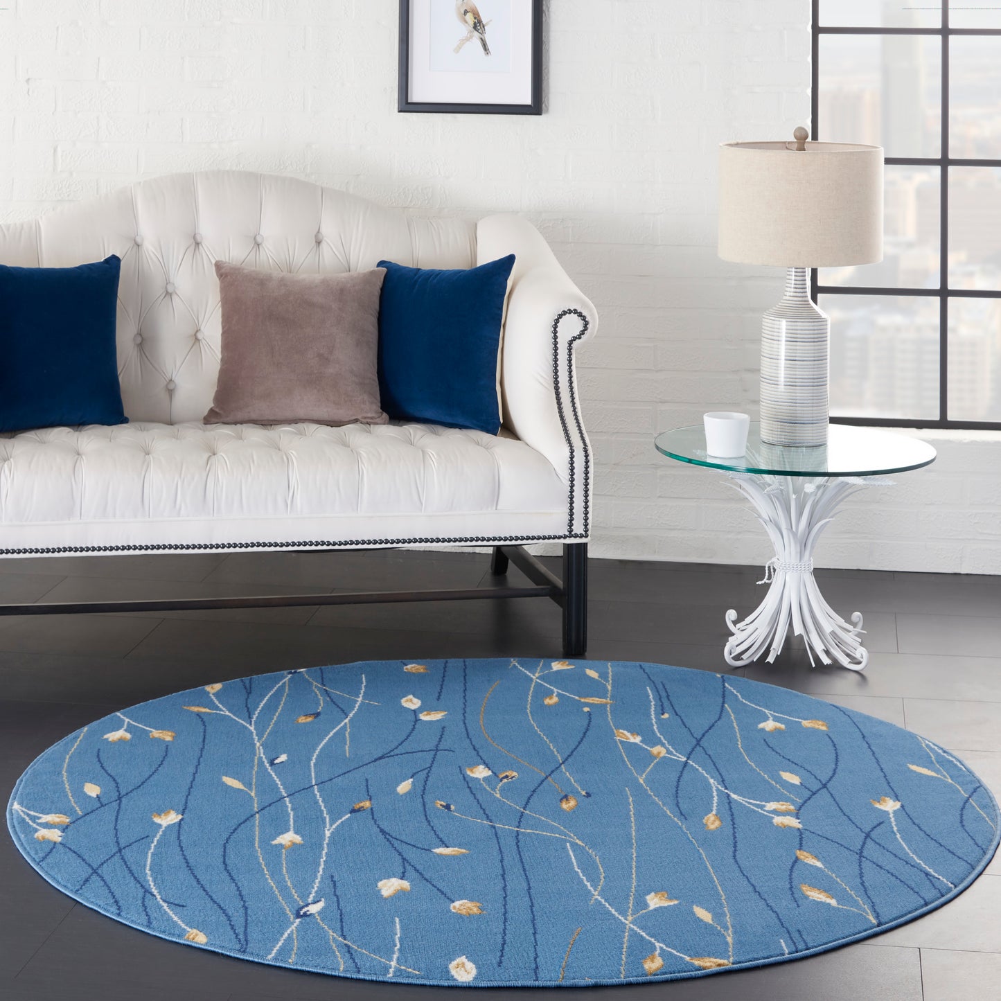 Round blue rug with ivory leaf vine pattern beside a white tufted sofa with nailhead trim, glass side table.