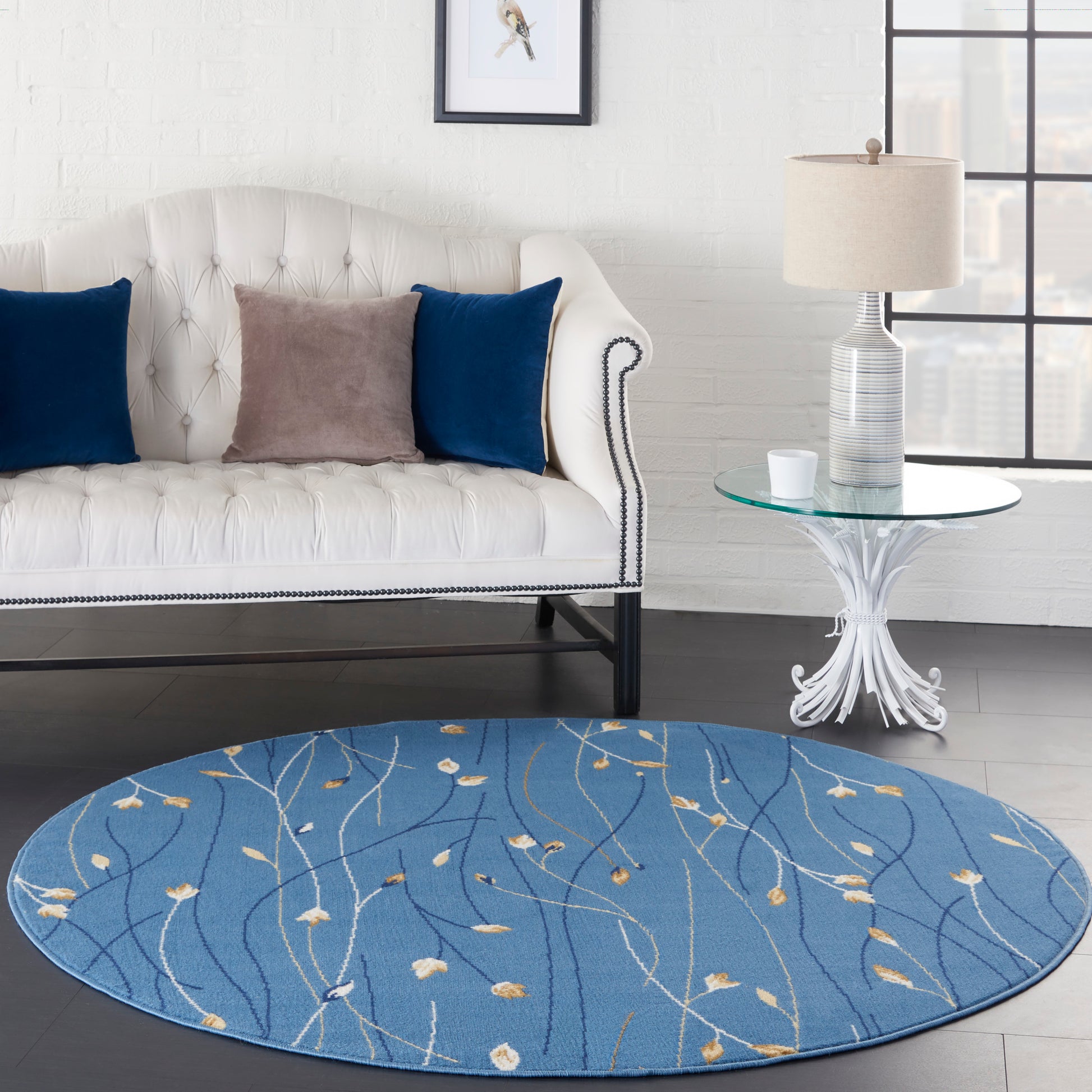 Round blue rug with ivory leaf vine pattern beside a white tufted sofa with nailhead trim, glass side table.