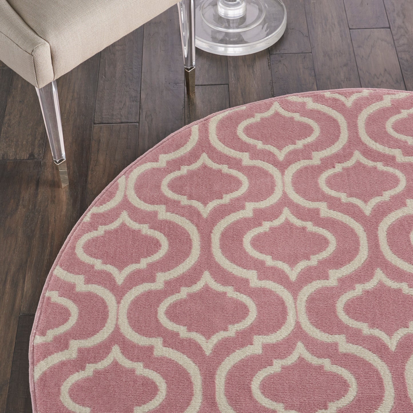 Round pink rug with cream trellis pattern on dark wood floor beside a beige chair.