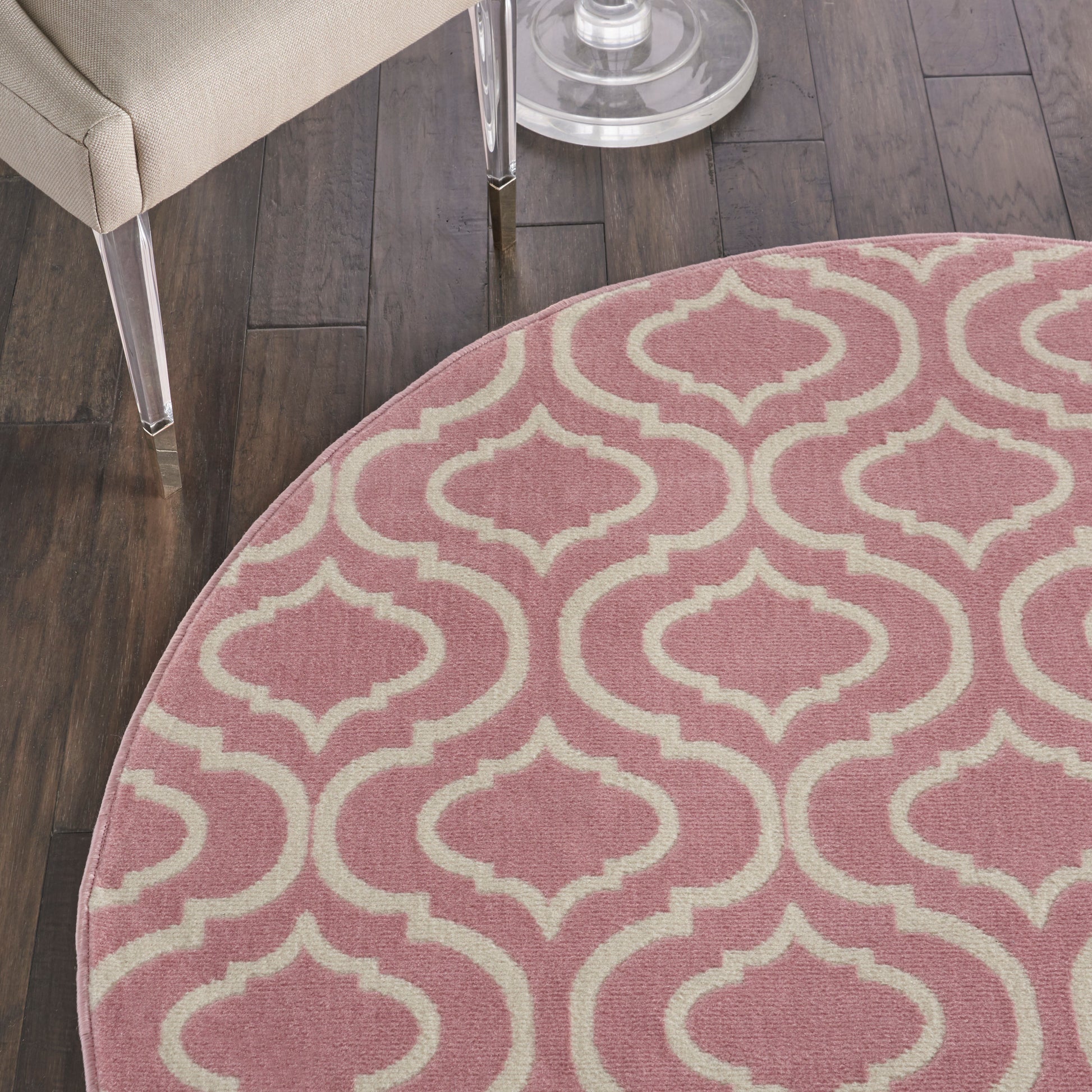 Round pink rug with cream trellis pattern on dark wood floor beside a beige chair.