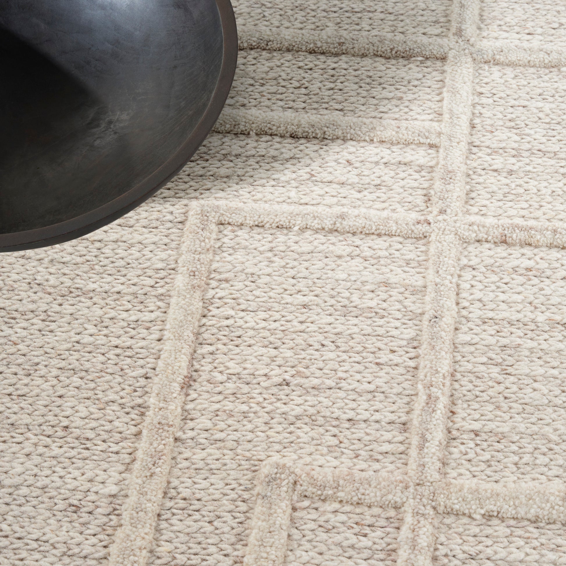 Beige textured woven rug with geometric raised line pattern; partial black round pan in the top-left.