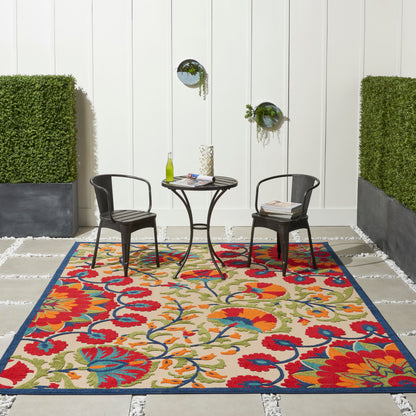 Outdoor scene: colorful floral rug with blue border, two black metal chairs, and a round table with a wine bottle and magazines.
