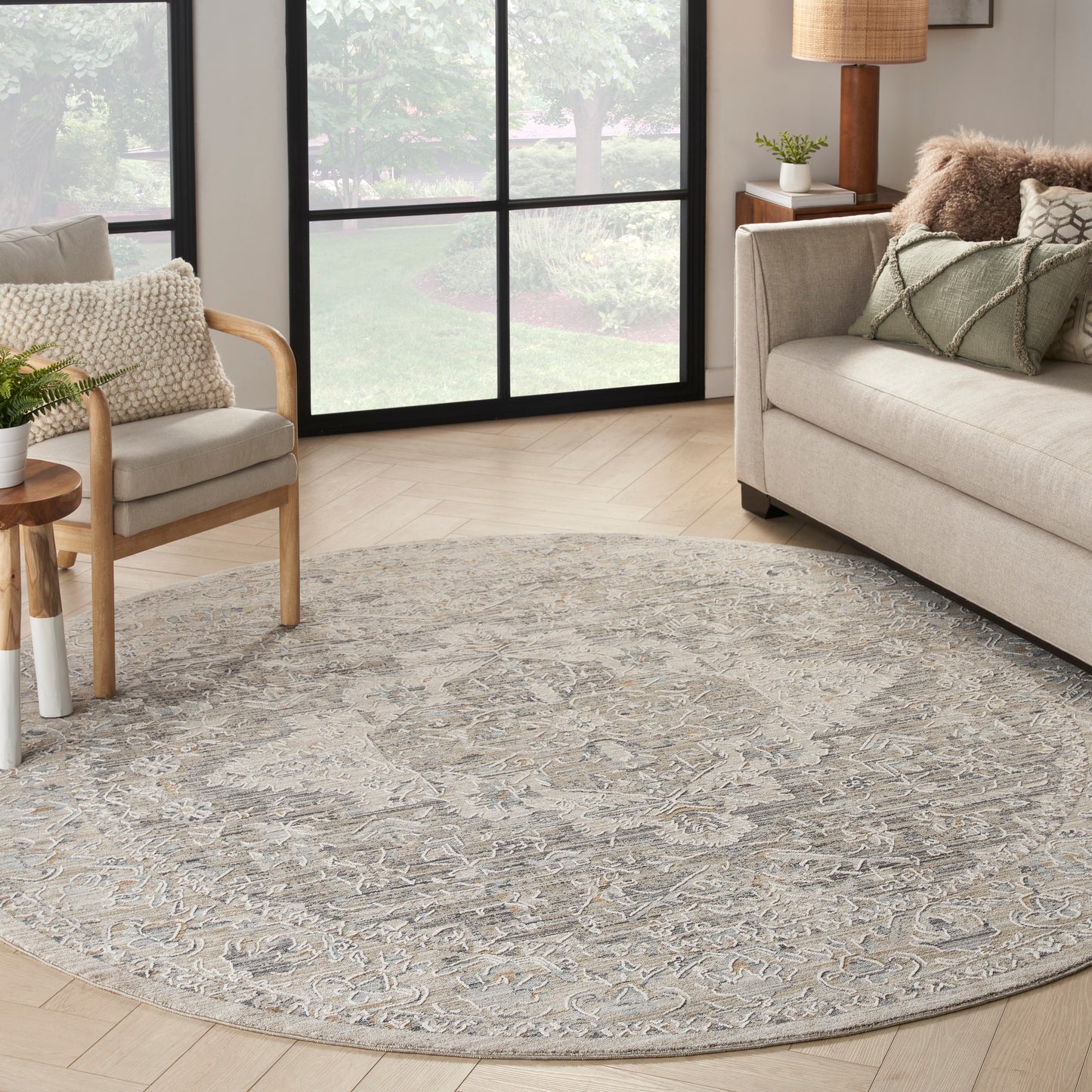 Round gray-beige ornate rug in a bright living room with beige sofa, wooden chair, and large grid windows.