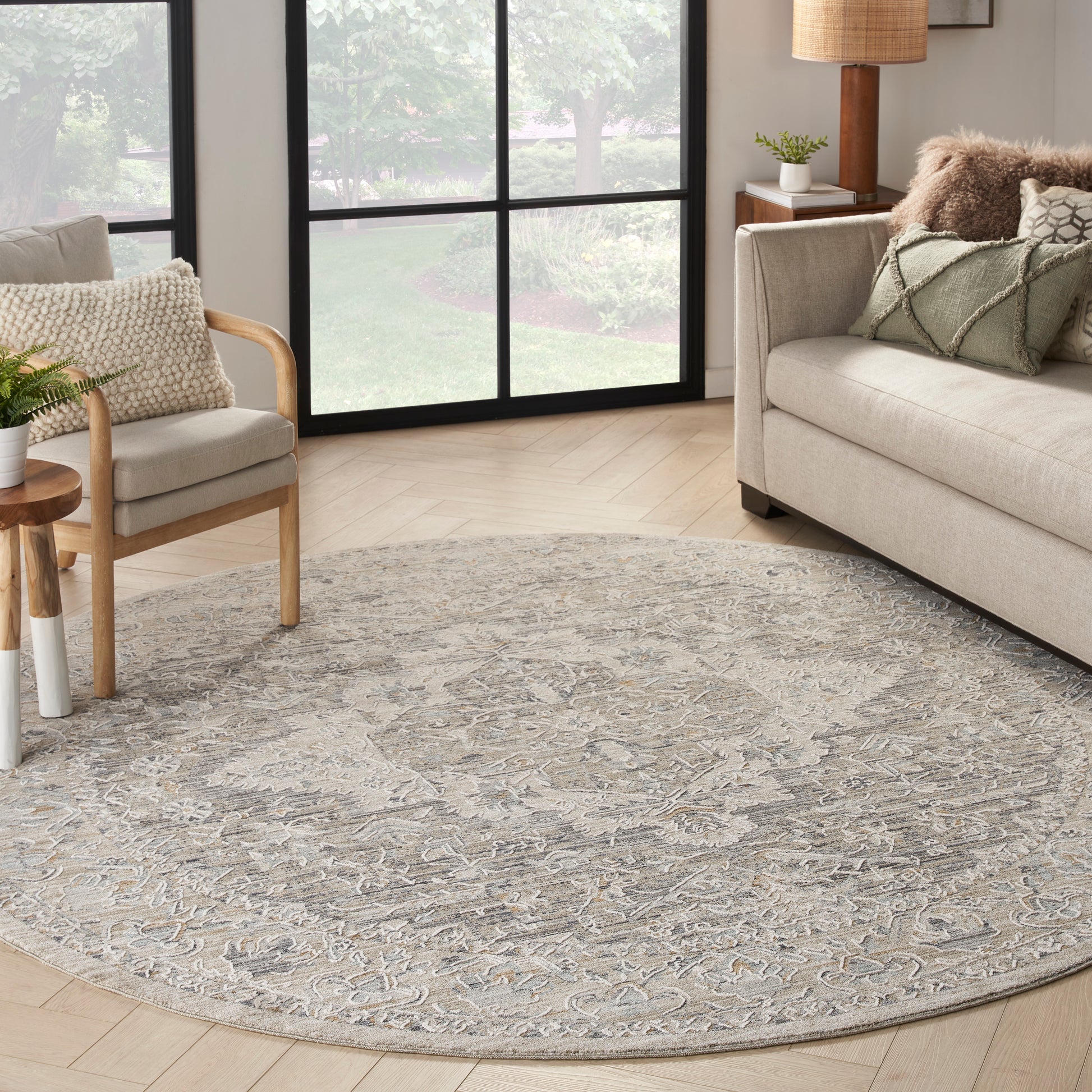 Round gray-beige ornate rug in a bright living room with beige sofa, wooden chair, and large grid windows.