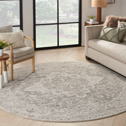 Round gray-beige ornate rug in a bright living room with beige sofa, wooden chair, and large grid windows.