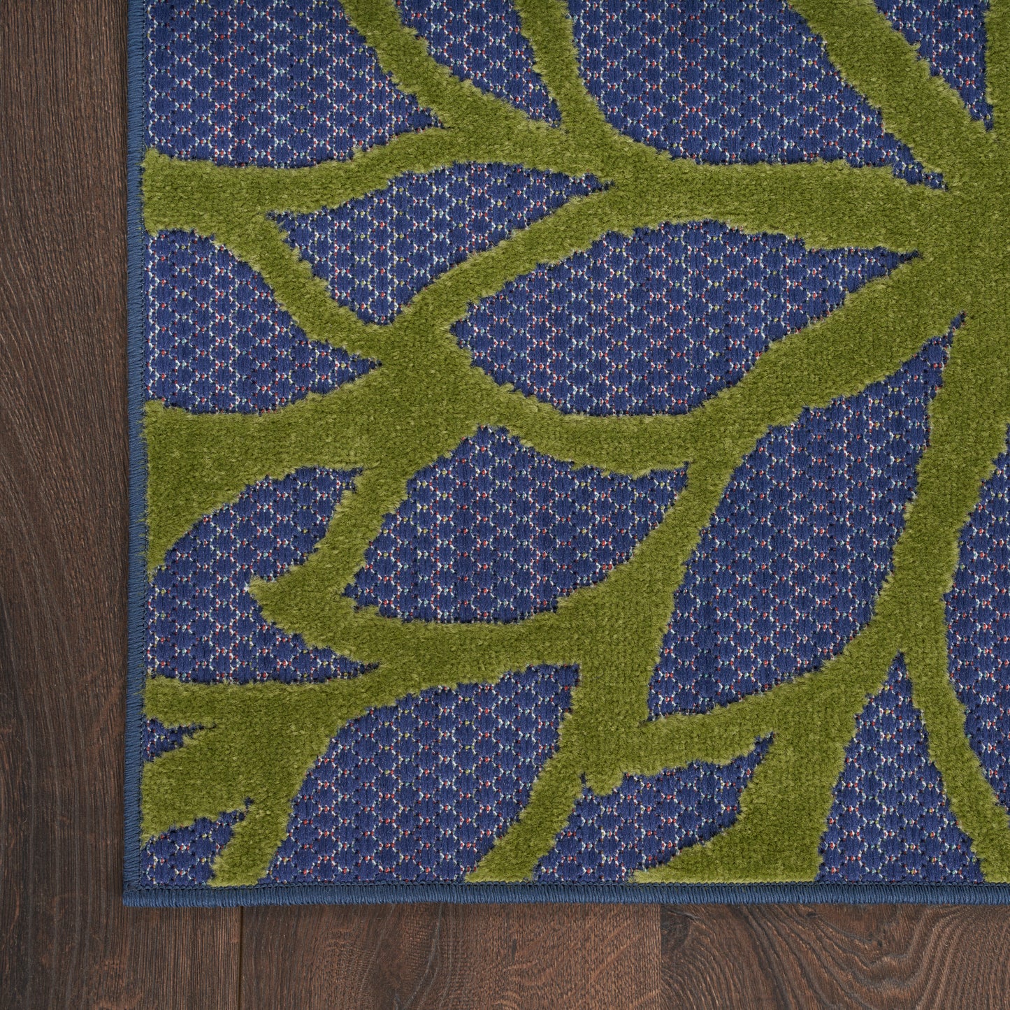 Blue tufted outdoor rug with an olive-green vine motif and blue border on dark wood floor.