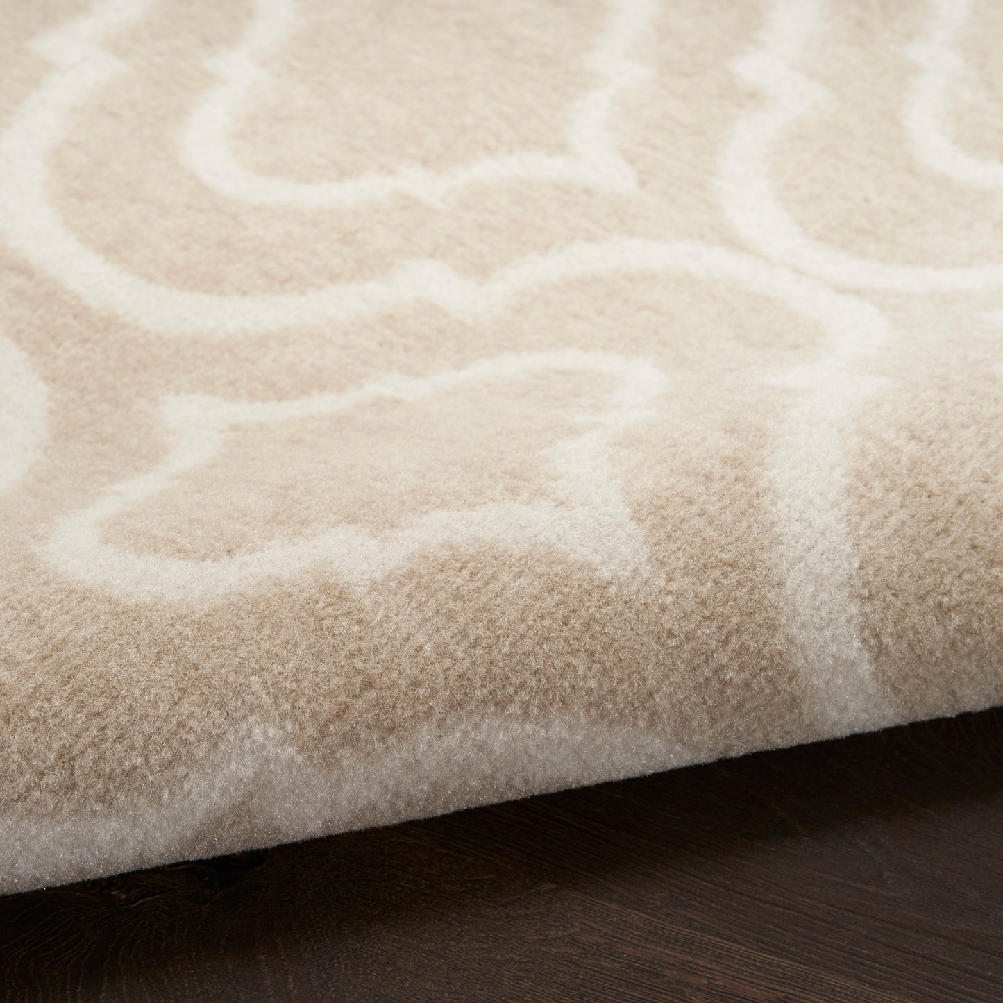 Beige plush area rug with white abstract pattern and soft pile, rolled edge on dark wood floor.