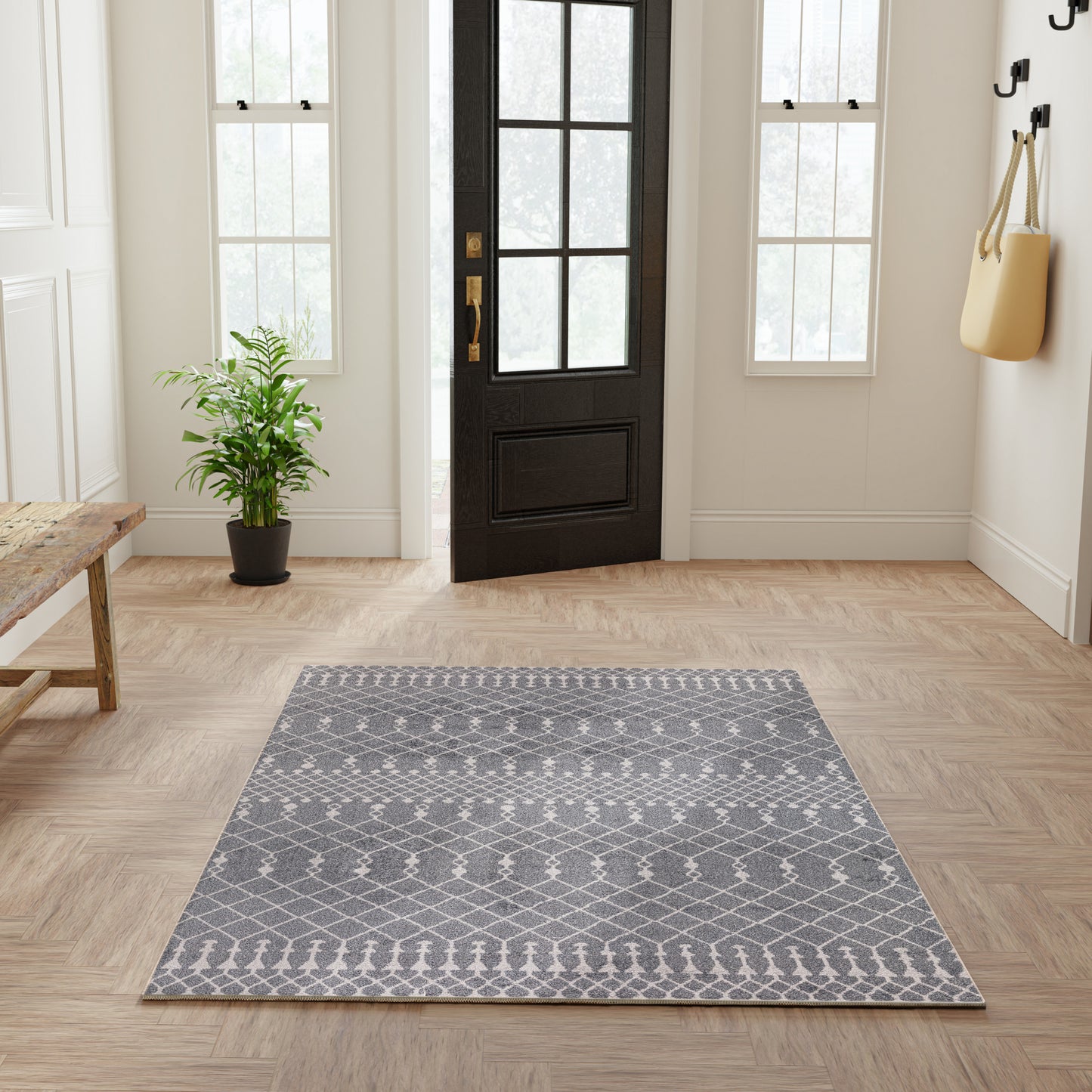 Gray geometric area rug on light wood entry floor; black door, potted plant, bench, and bag on hooks.