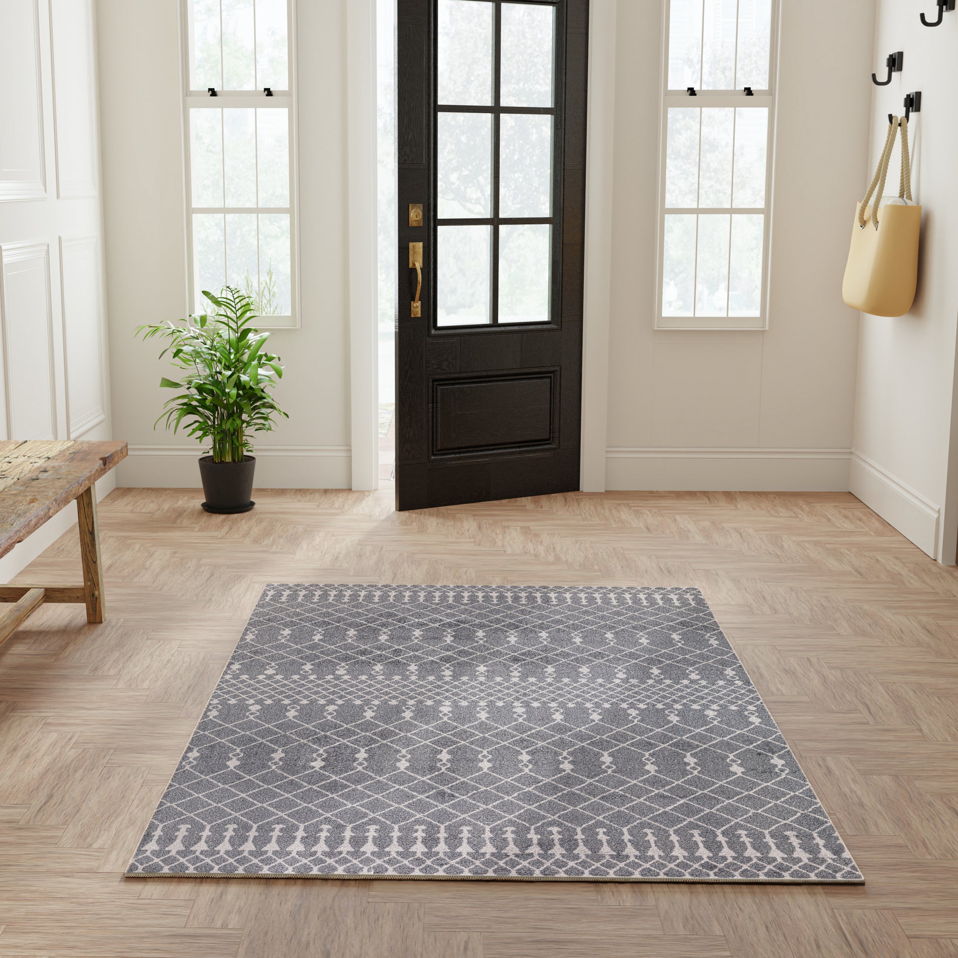Gray geometric area rug on light wood entry floor; black door, potted plant, bench, and bag on hooks.