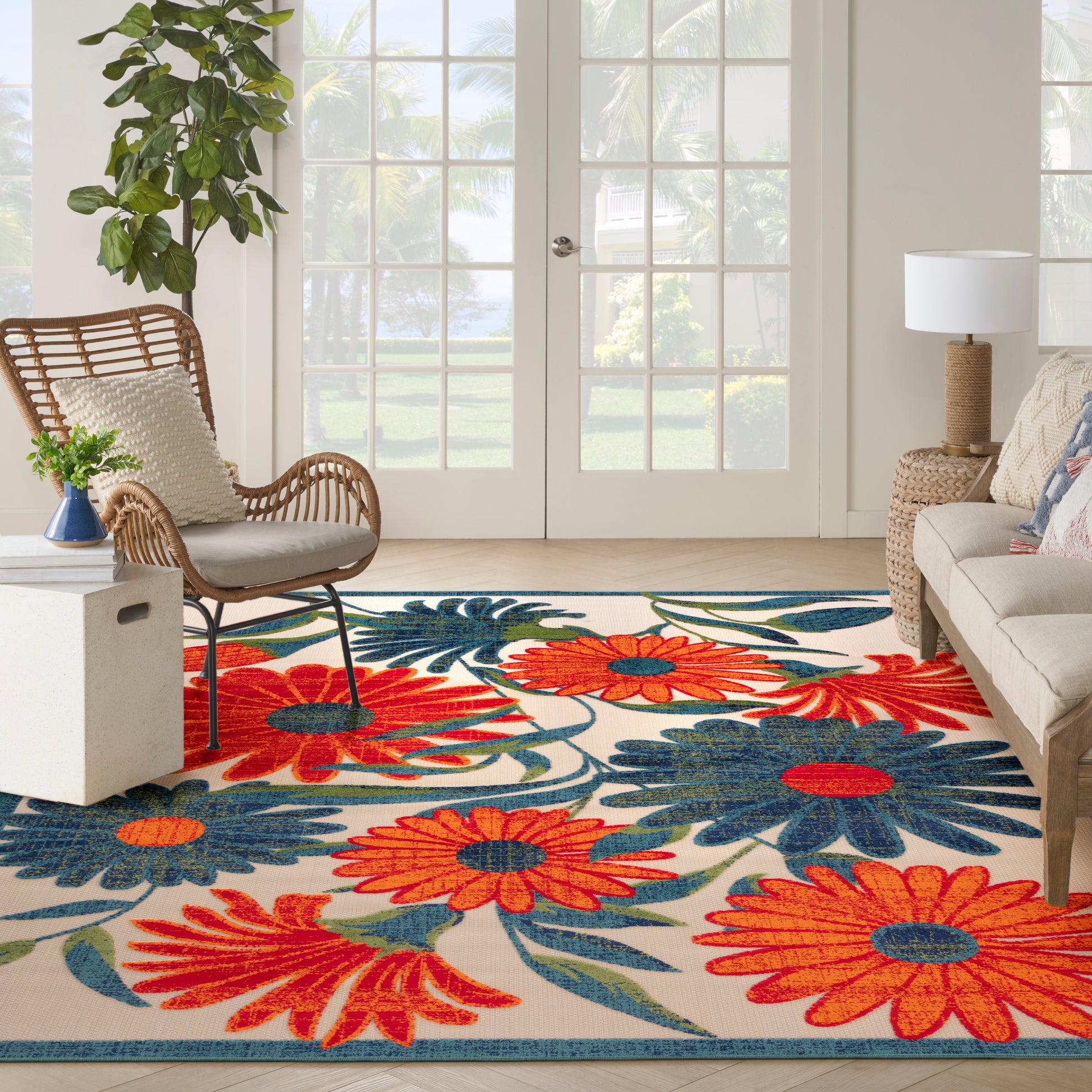 Bright outdoor living area with a bold tropical floral rug in red, orange and blue; wicker chair, beige sofa, and a potted plant.
