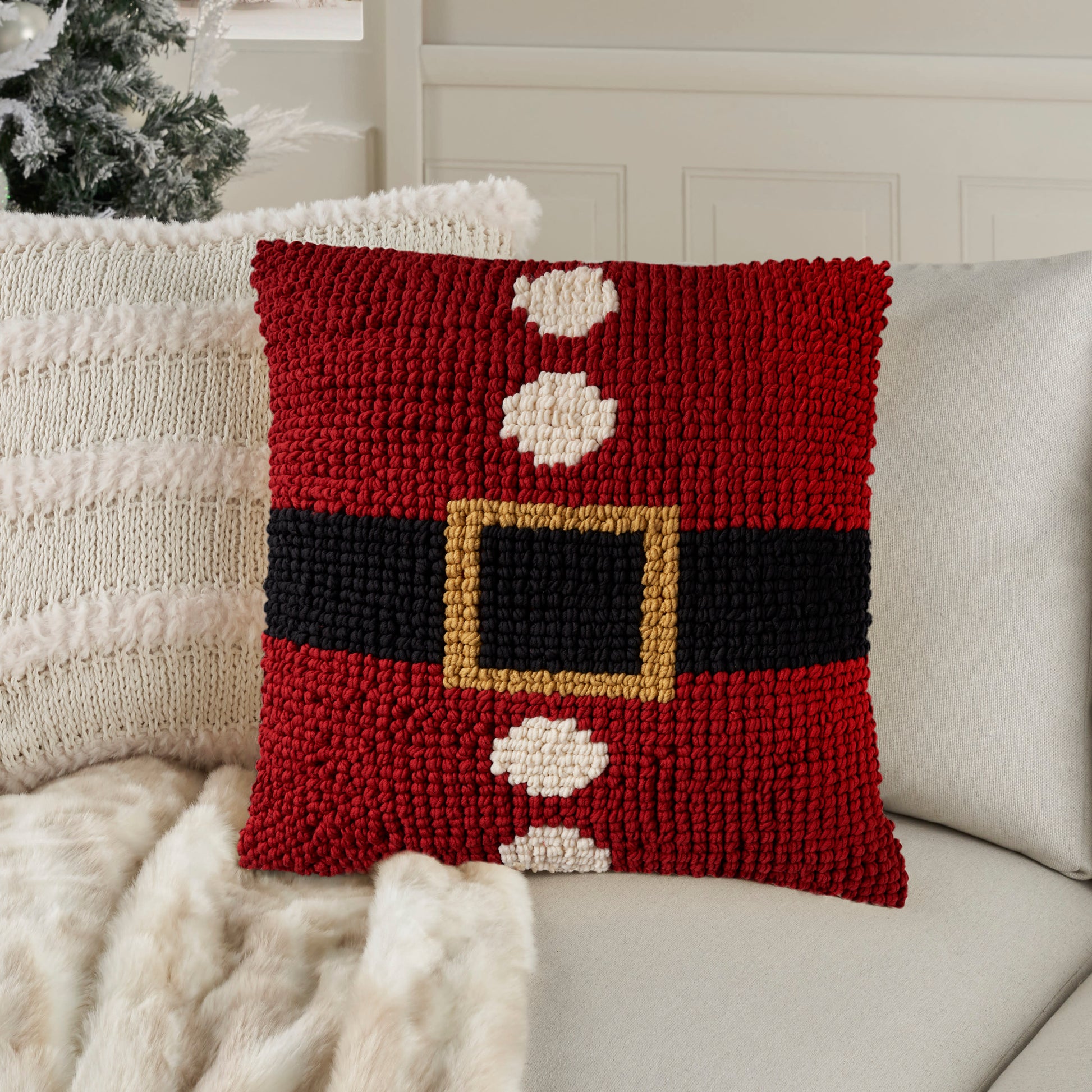 Red pillow with Santa belt motif: black belt, gold buckle, white circles along belt, on light sofa.