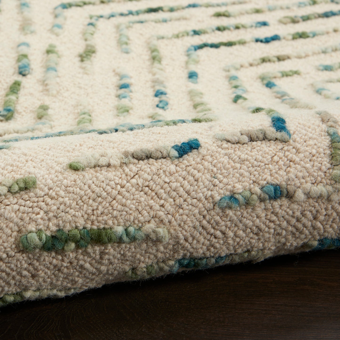 Close-up of a beige shag rug with blue & green yarn accents in a linear geometric pattern on a dark wood floor.