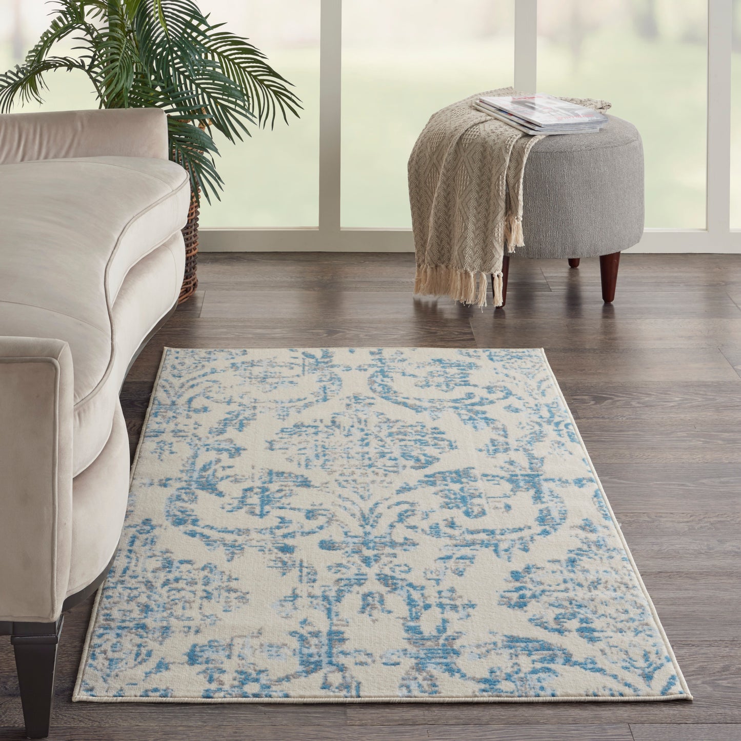 Cream and blue patterned area rug on dark wood floor in a bright living room with a beige sofa and round gray ottoman.