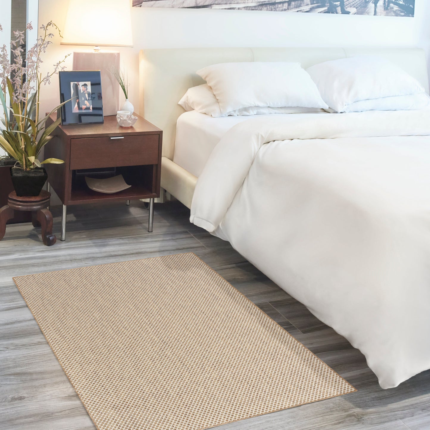 White bed with beige woven rug, dark wood nightstand, plant and framed photo in a bright cozy bedroom.