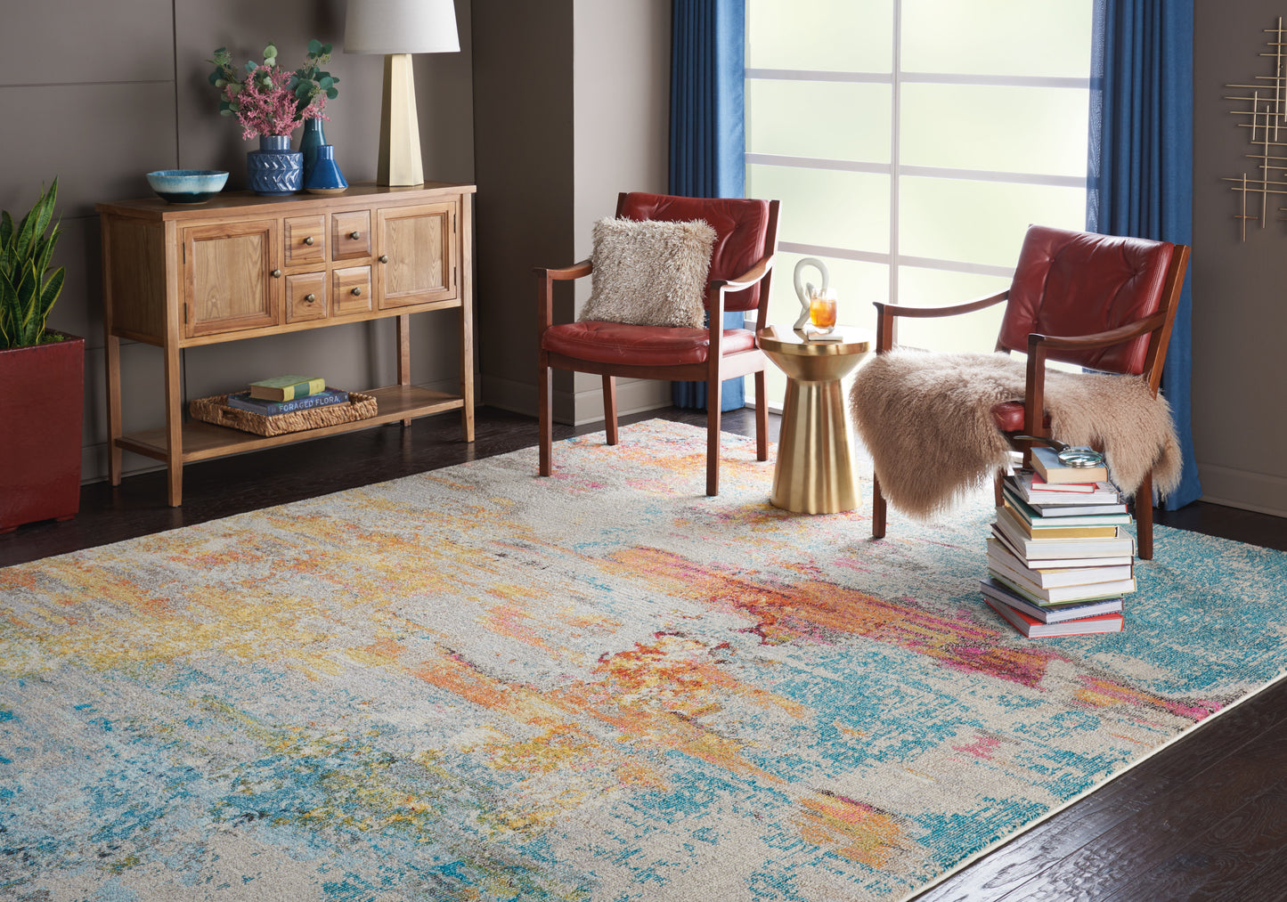 Large abstract multicolor rug in blues and yellows; red leather chairs with fur throws, wooden console, gold side table.