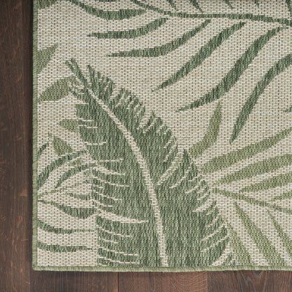 Rectangular woven rug with a botanical palm leaf pattern in green on beige, textured weave, edge bound, on dark wood floor.