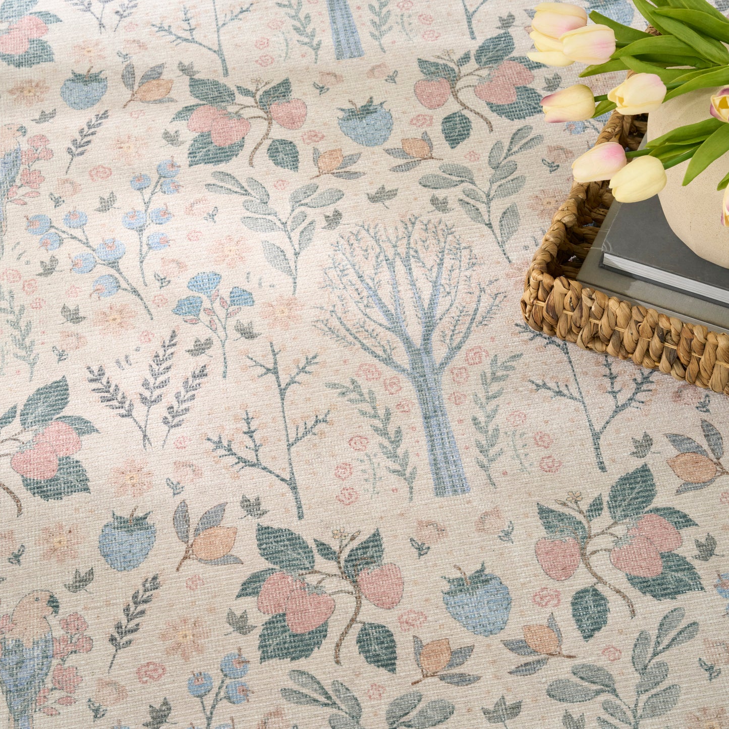 Pastel botanical rug with tree and leaf motifs in blue, pink, and sage; woven basket with books and a vase of tulips.