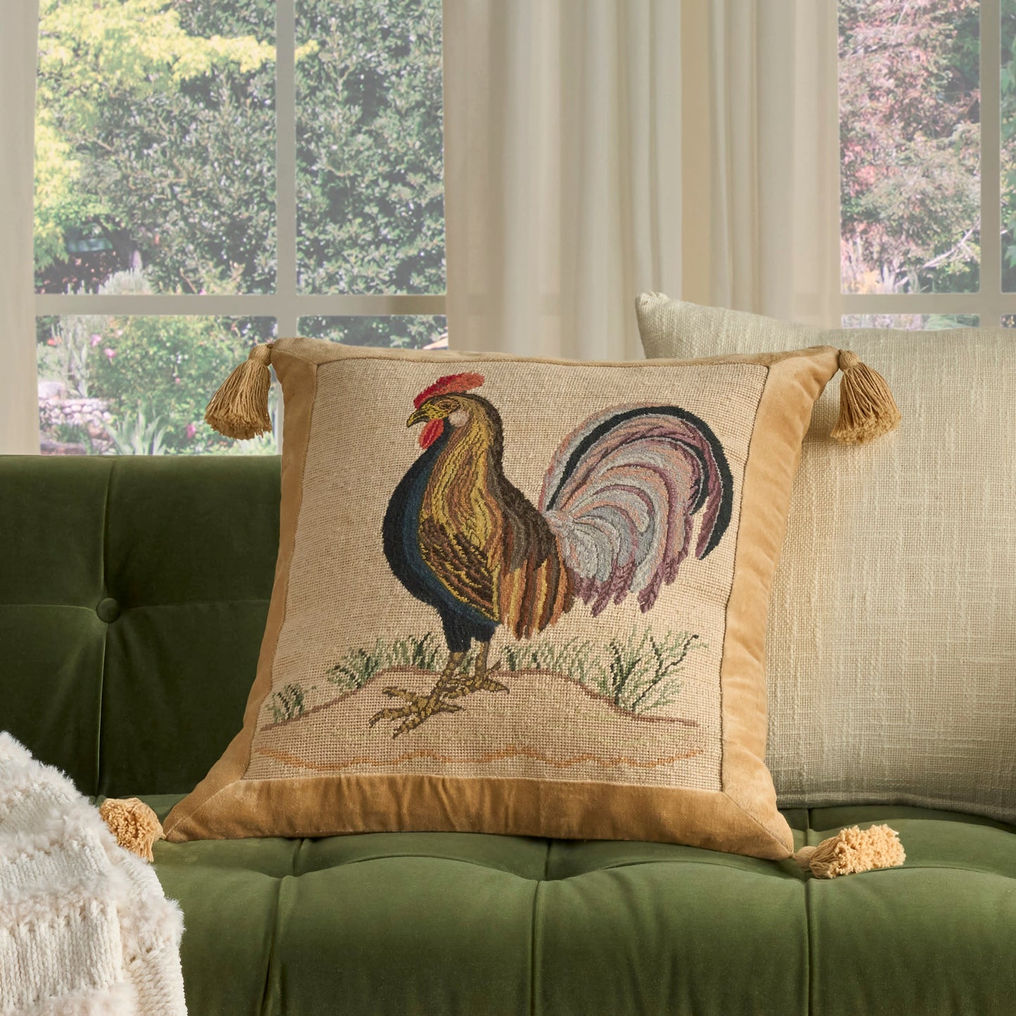 Beige decorative throw pillow with rooster embroidery, gold tassels on corners, on green tufted sofa in sunlit living room.