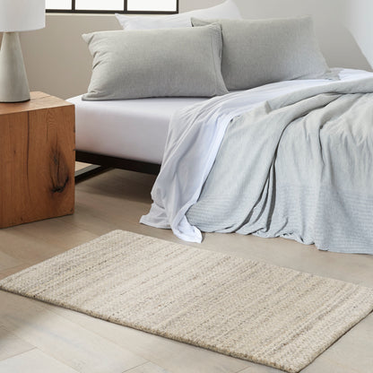 Neutral bedroom with a low bed, light gray bedding, white sheets, beige woven rug, and wooden nightstand with lamp.