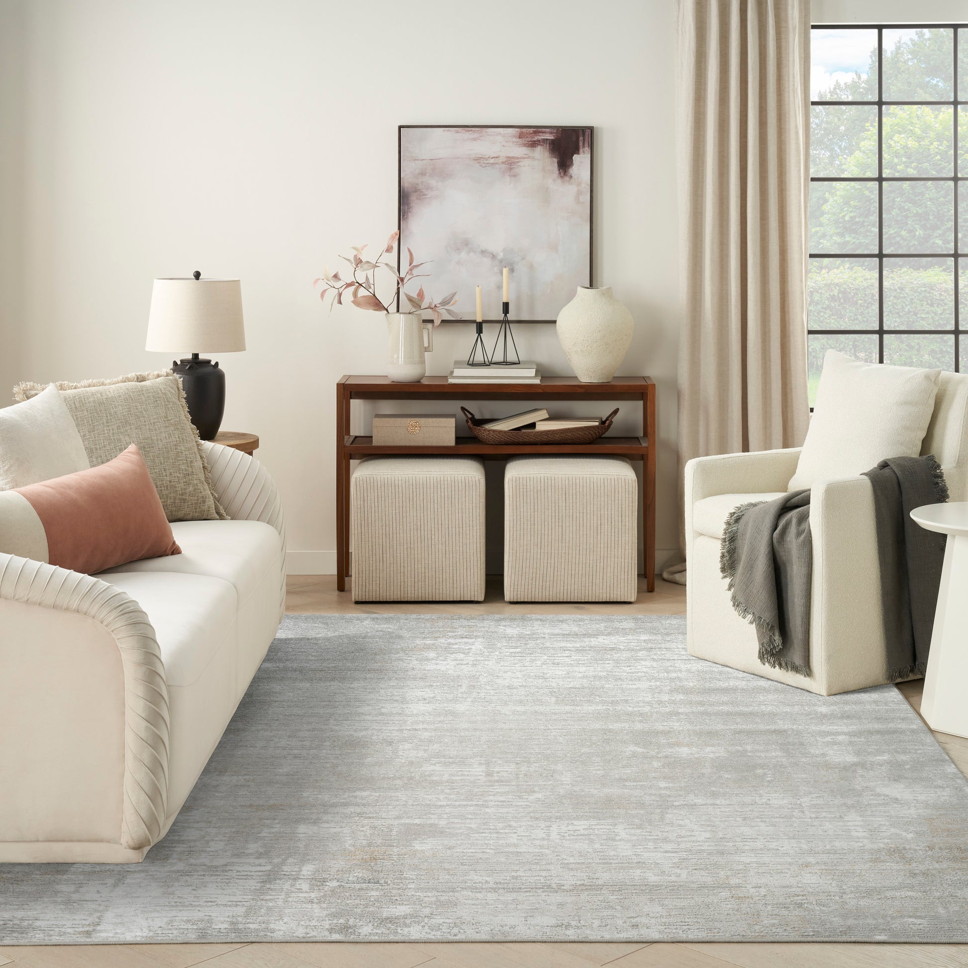 Neutral living room with cream sofa, blush pillow, wooden console, framed abstract art, two beige ottomans, light blue rug.