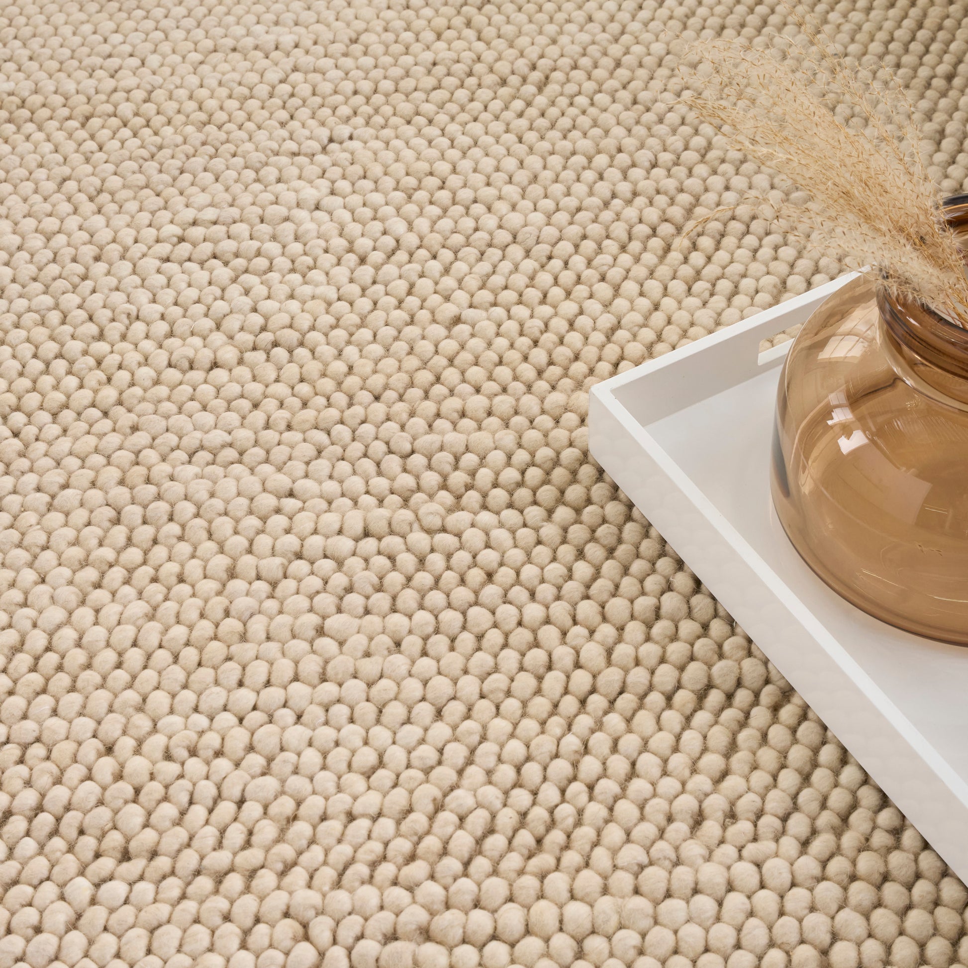 Beige textured rug with a white tray holding an amber glass vase and dried grasses.