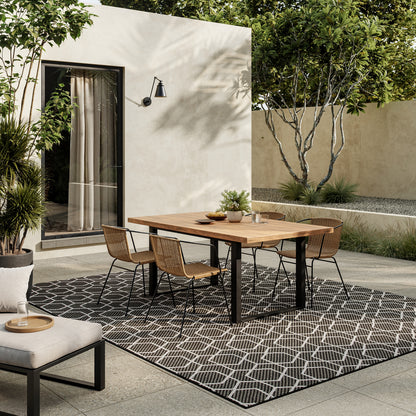 Modern outdoor dining: wood table, black legs, wicker chairs, geometric rug on patio beside a beige wall.