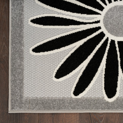 Gray rug with a large black petal flower outlined in ivory, textured border, on dark wood floor.