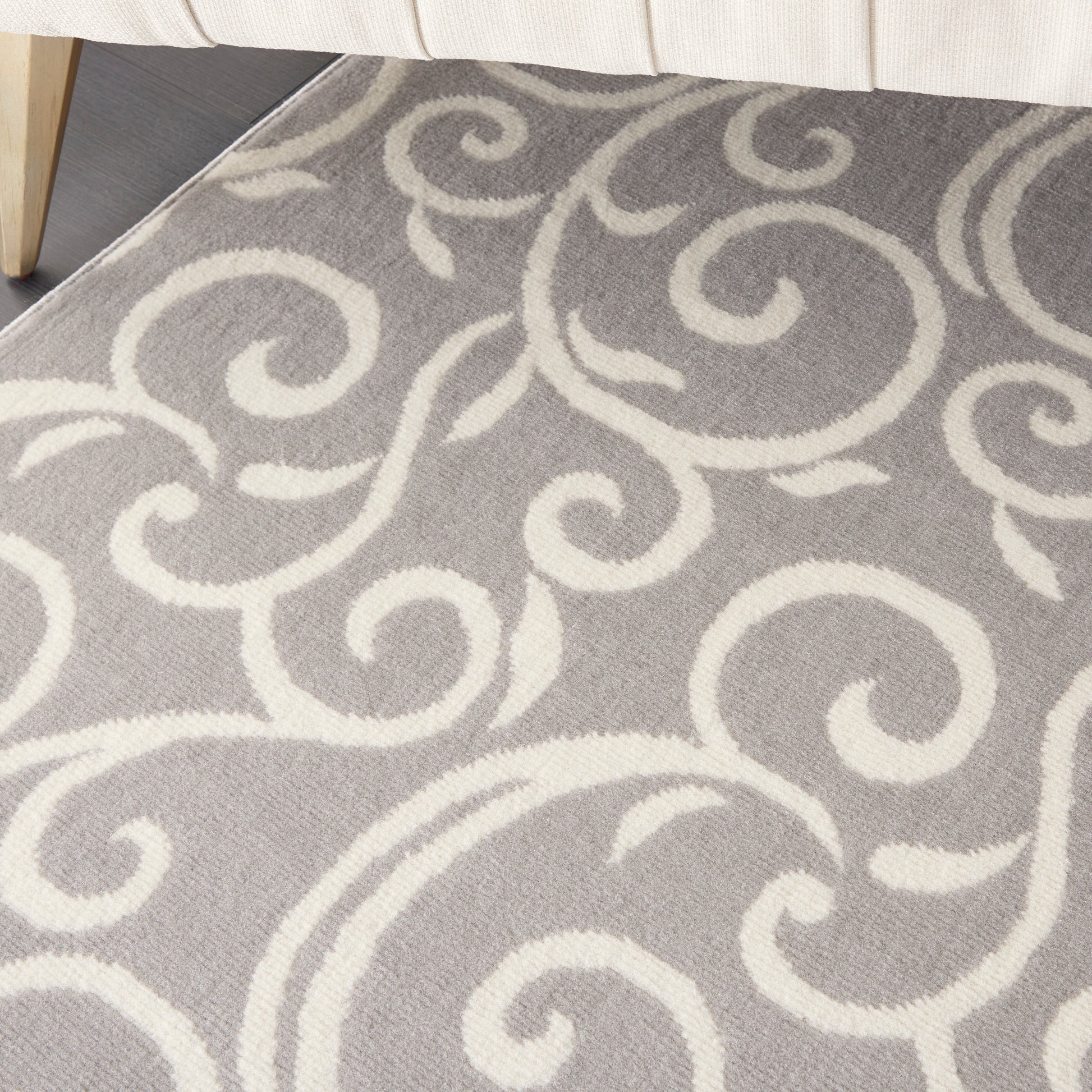 Soft gray rug with white scroll-pattern design; partial beige sofa leg visible.