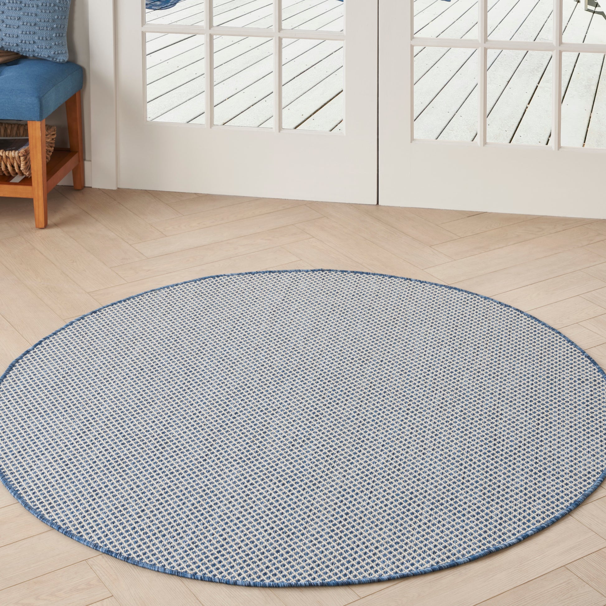 Round blue-gray woven rug with blue border on light wood floor beside glass doors.