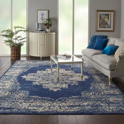Coastal living room with navy distressed area rug, glass-top coffee table, cream sofa with blue cushions, curved white cabinet, plants and framed art.