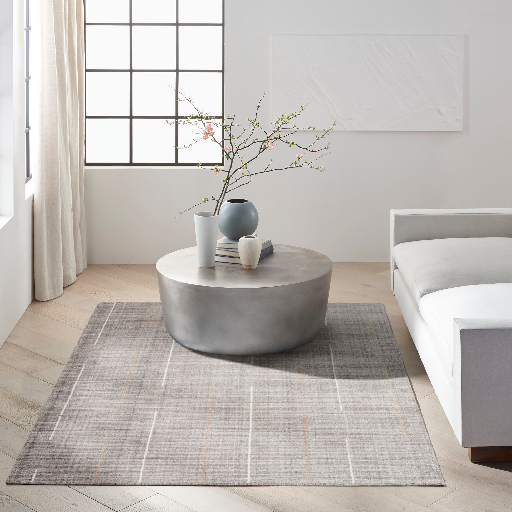 Gray grid-pattern rug in a minimalist living room with a white sofa, round coffee table, vase with branches, and books.