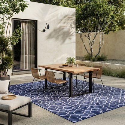 Outdoor dining set with wooden-top table and black metal legs, wicker chairs on a blue geometric rug beside beige patio wall