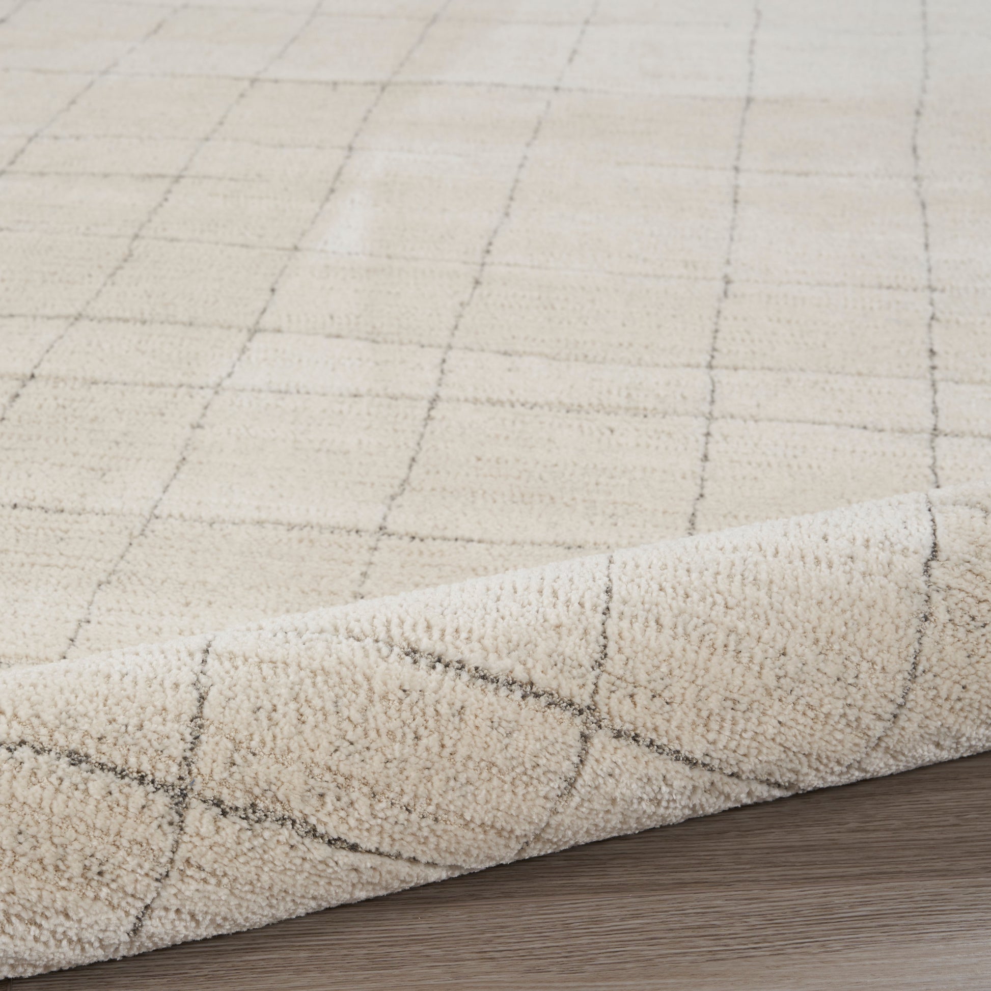 Beige rug with subtle dark grid pattern; short pile texture, rolled corner on wooden floor.