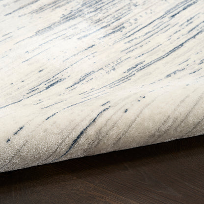 Cream area rug with blue-gray abstract brushstroke pattern and soft plush pile on dark wood floor.