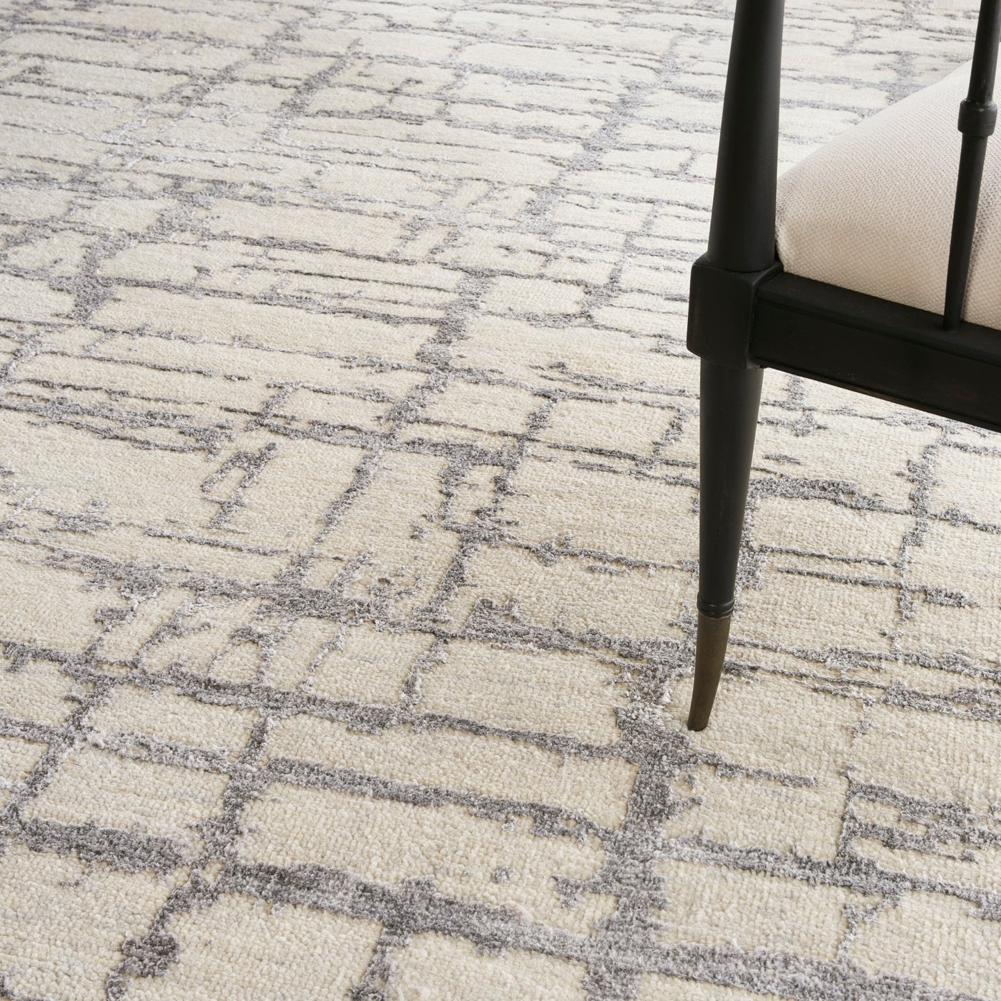 Beige textured rug with a gray geometric lattice pattern, paired with a black chair leg and light beige upholstery.