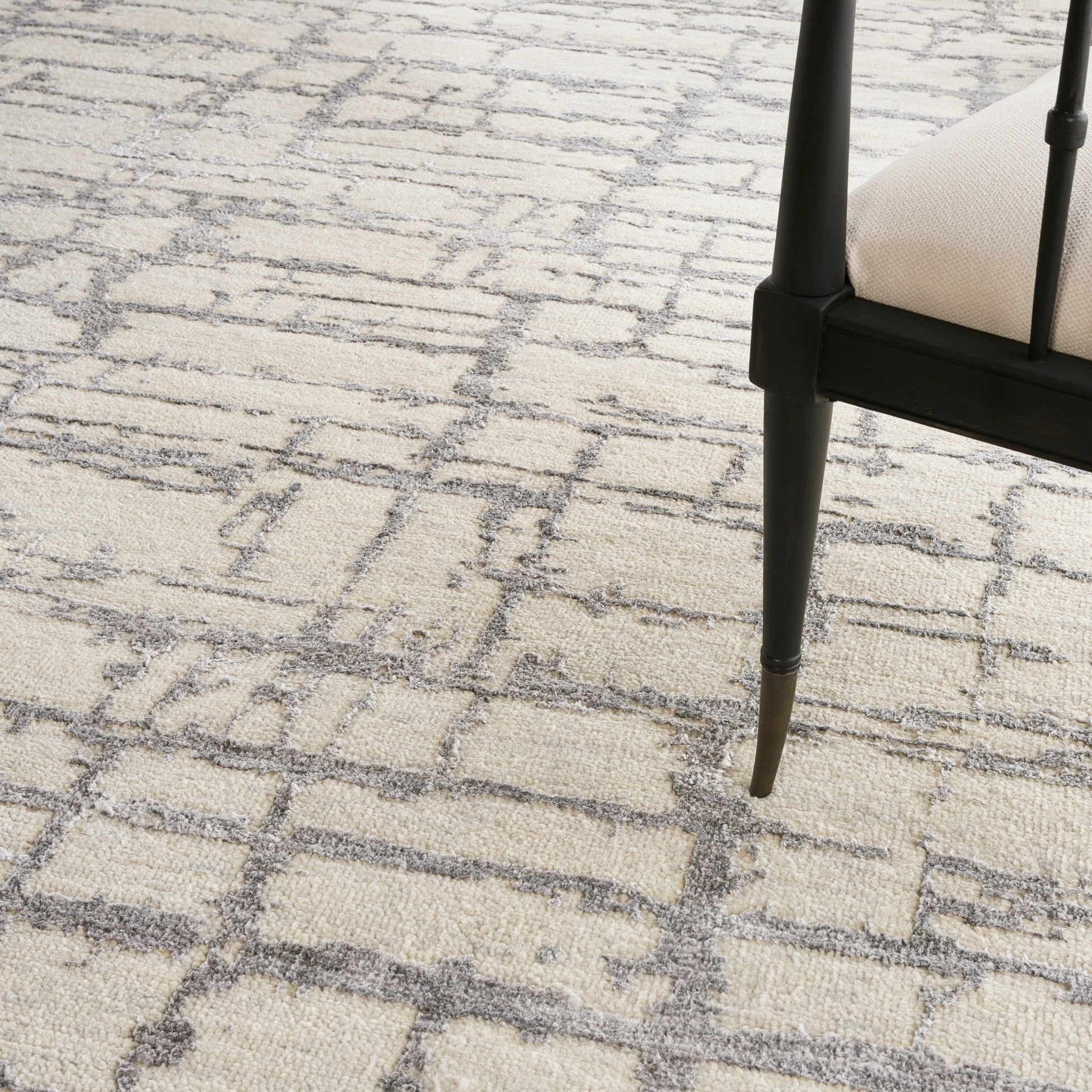 Beige textured rug with a gray geometric lattice pattern, paired with a black chair leg and light beige upholstery.