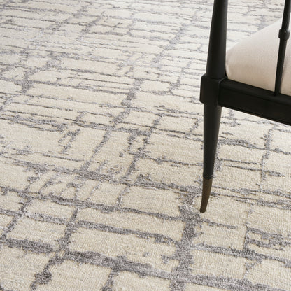 Beige textured rug with a gray geometric lattice pattern, paired with a black chair leg and light beige upholstery.